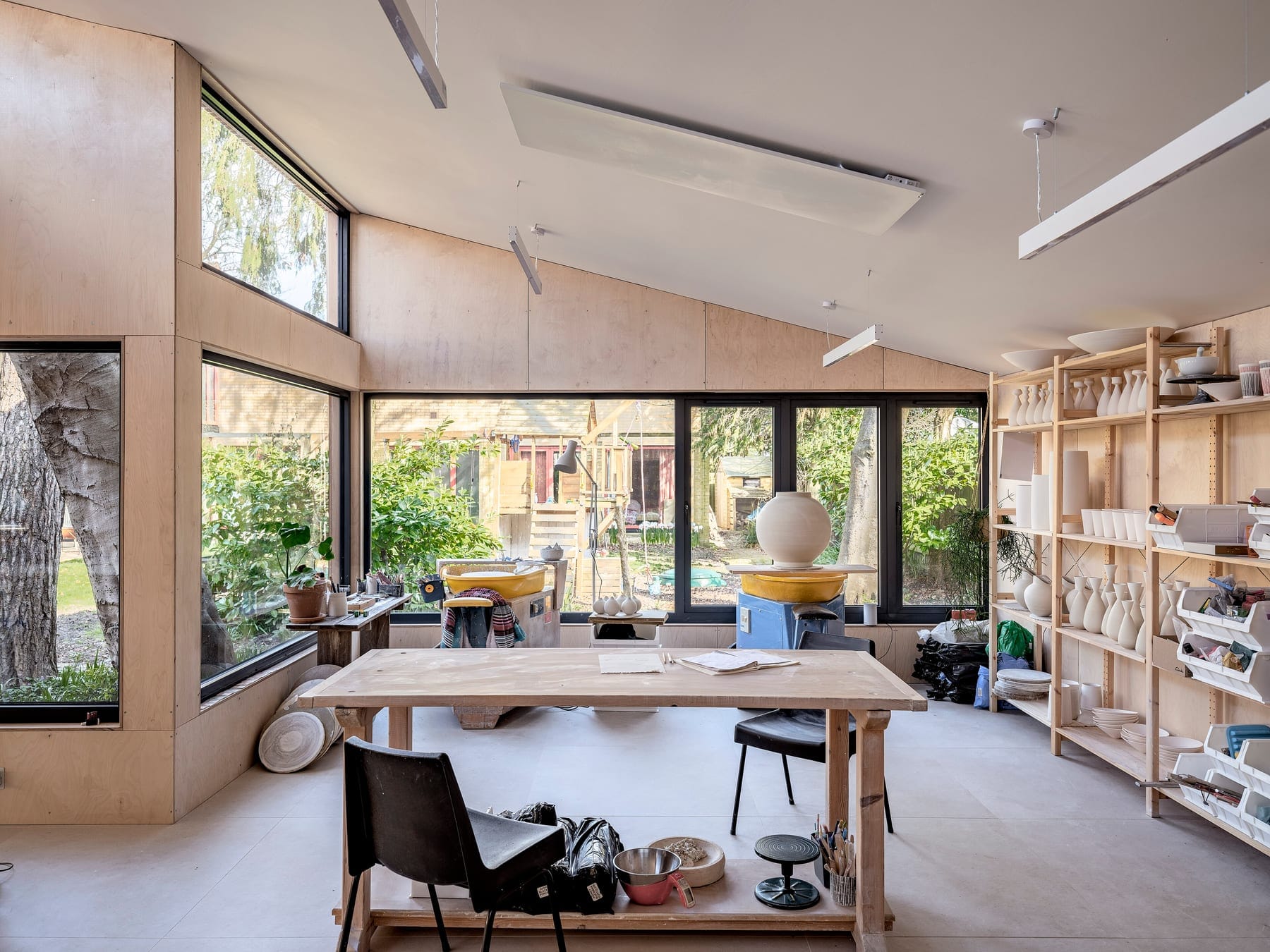 A bright pottery studio with a wheel, a long worktable and shelves of white ceramic forms, seen through large windows onto a garden. Generic studio photograph; it does not depict a listed school.